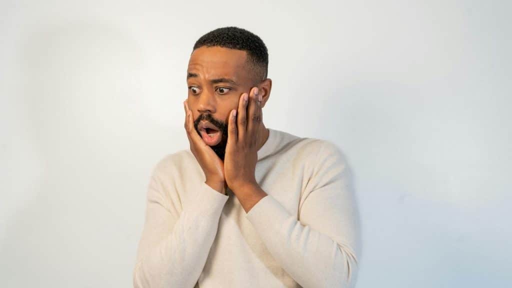 A shocked man holds his face with wide eyes against a plain background.