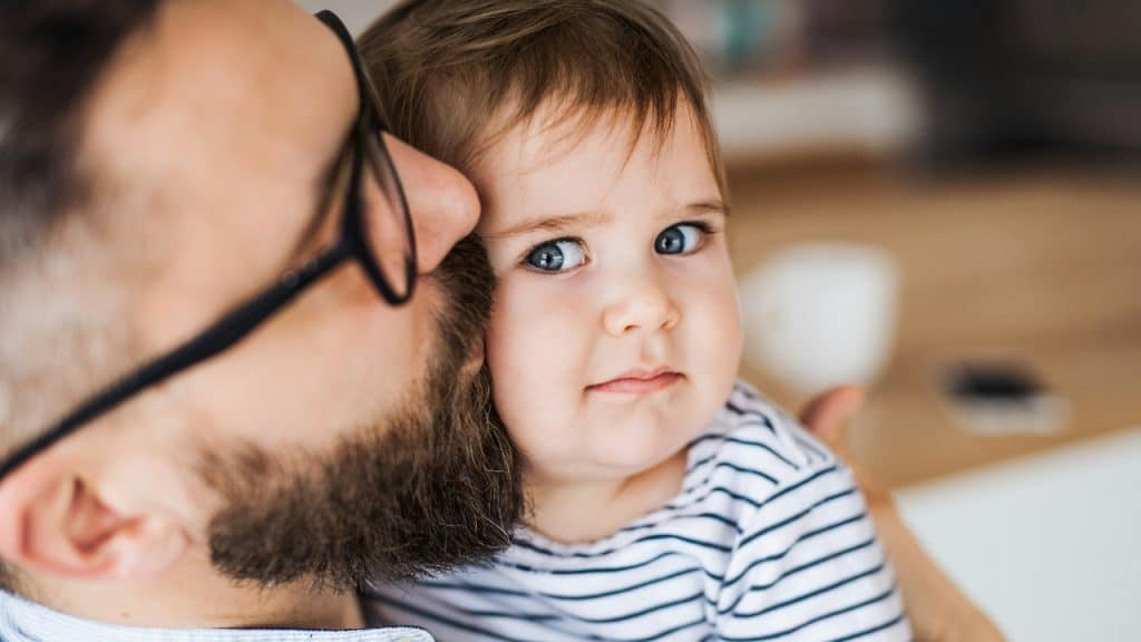 A bearded man wearing glasses kisses a young child on the cheek in a close-up.