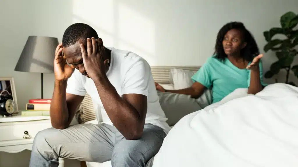A man sits on a bed holding his head while a woman gestures nearby.