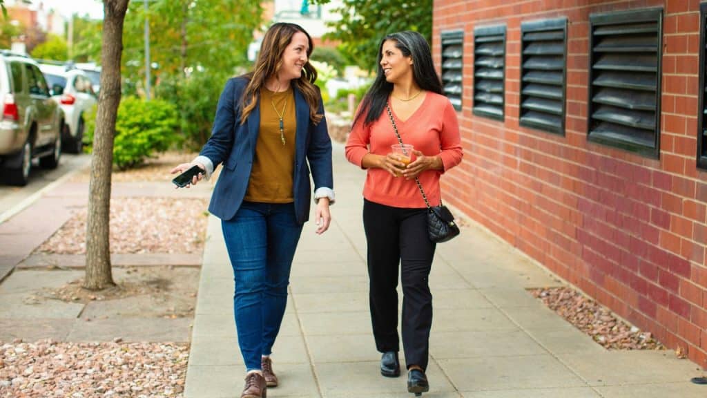 Two women walk together on a sidewalk past a brick building and a line of cars.