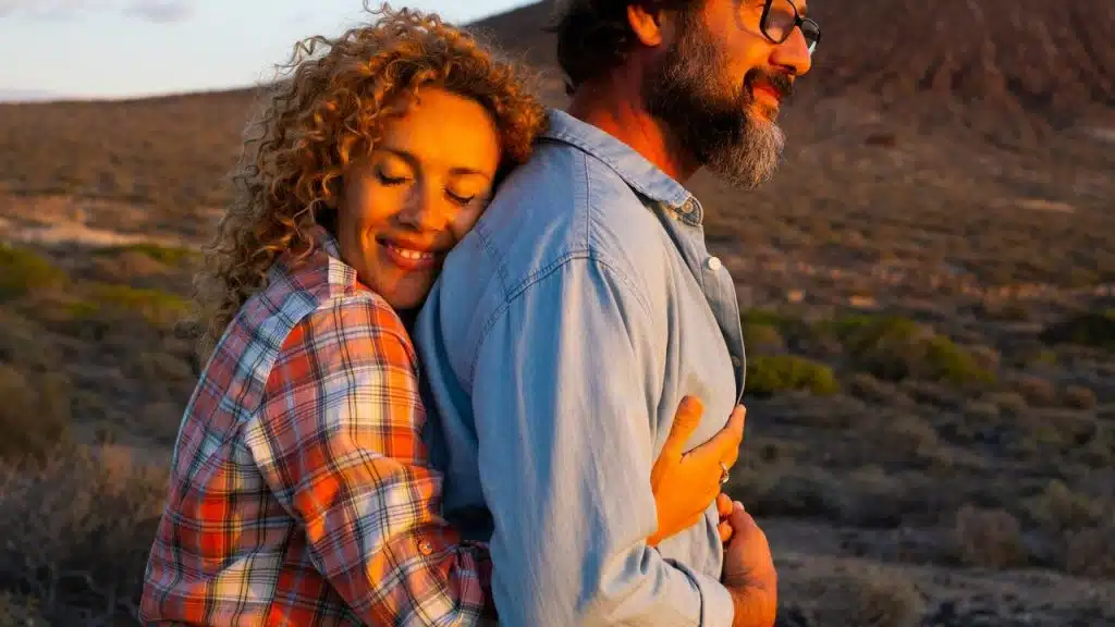 A woman hugging a man from behind outdoors at sunset.