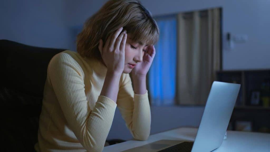 A woman holding her head while sitting at a laptop at night.