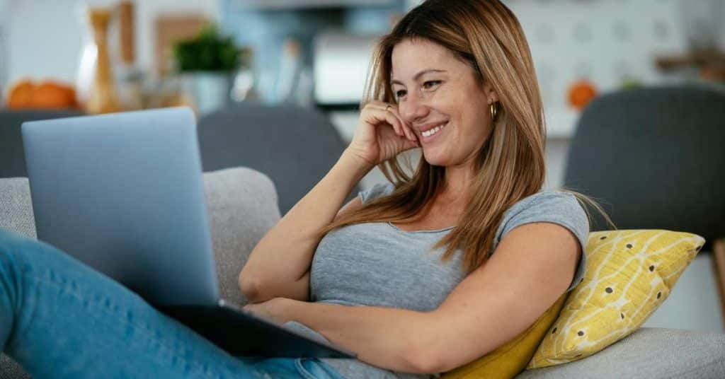 A woman smiling while using a laptop on a couch.