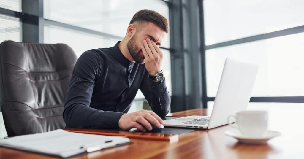 A man sitting at a desk rubbing his eyes while working on a laptop.