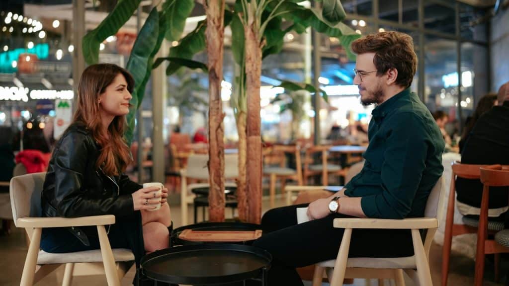 A man and woman sit across from each other at a table with white mugs.