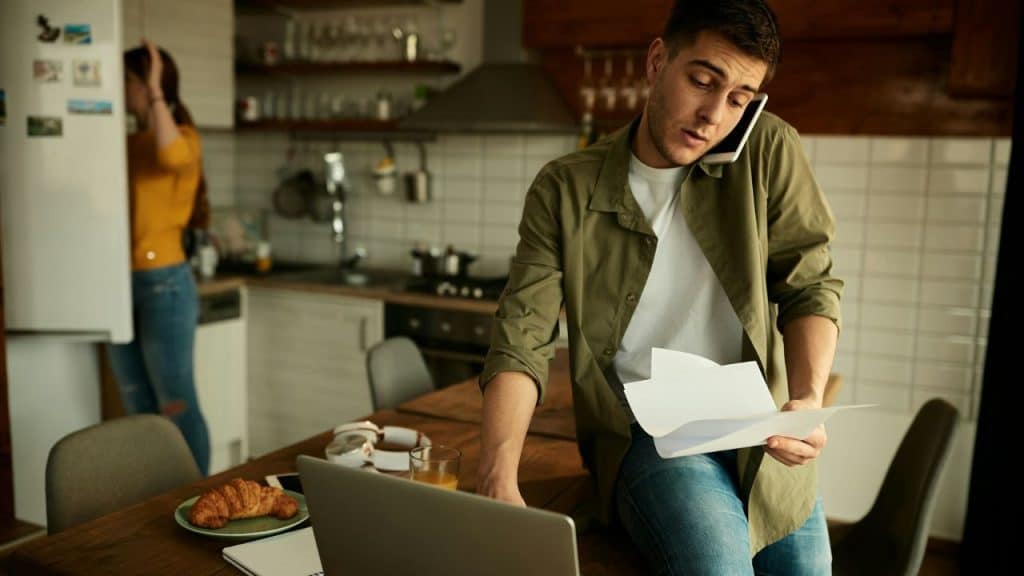 A man speaks on a phone while holding papers near a laptop in a kitchen.