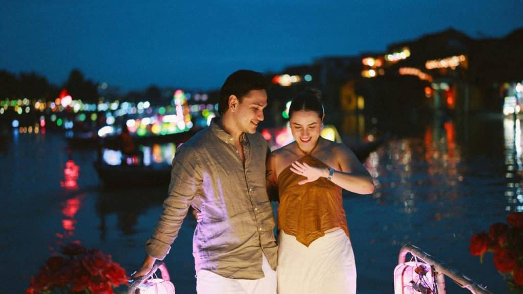A couple walking together at night by a river lit with colorful lanterns.