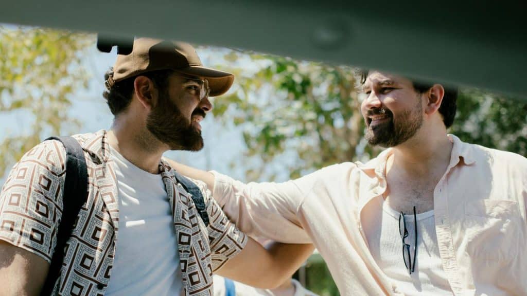 A pair of men smiling at each other outdoors with an arm around a shoulder.