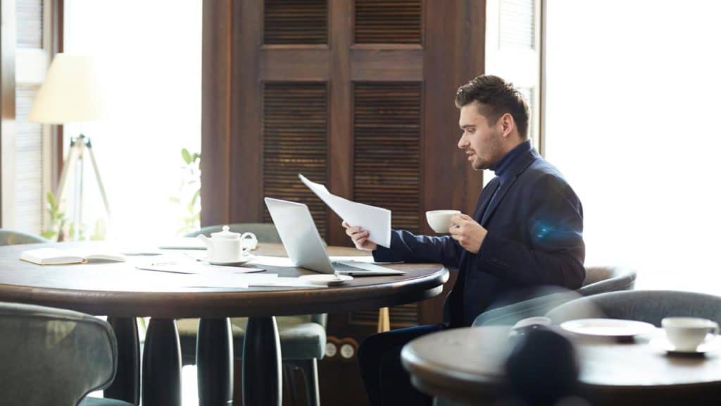 A man sitting at a table, reviewing papers while holding a cup of coffee beside a laptop.