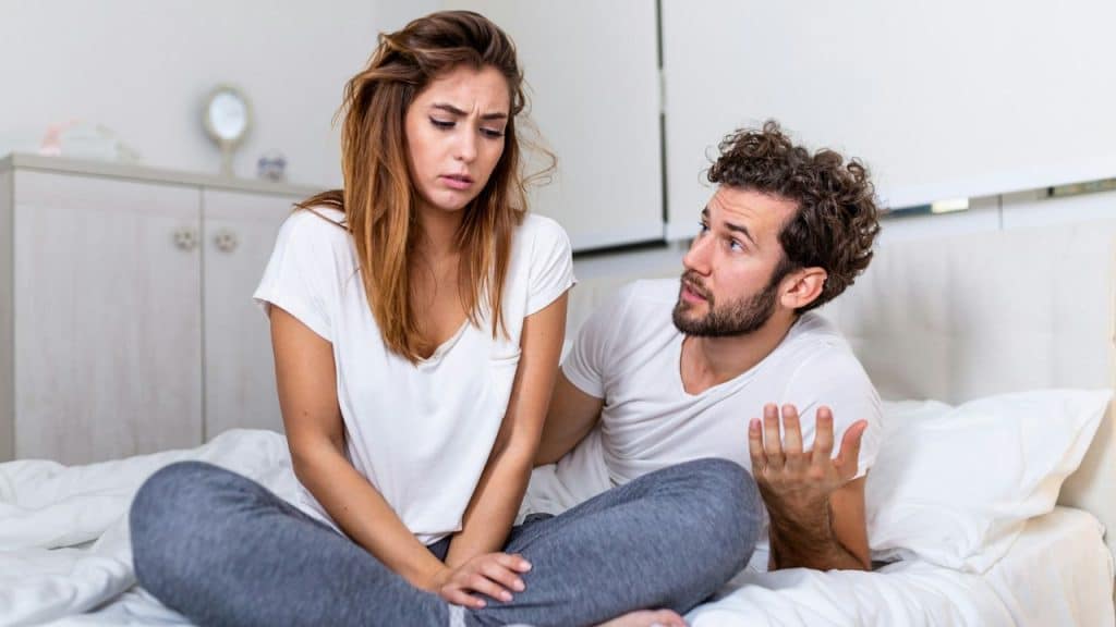 A man gestures while talking to a woman sitting on a bed with a concerned expression.