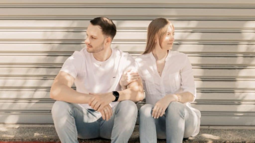 A man and woman in white shirts sitting outdoors against a metallic slatted shutter background.