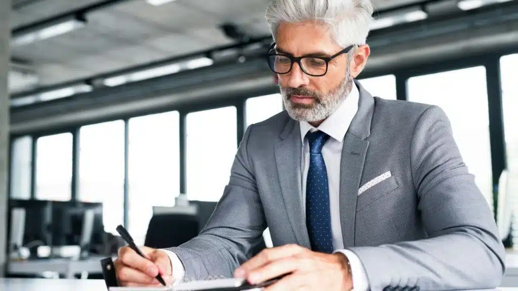 A grey-haired man in a suit and glasses writes in a notebook at a desk.