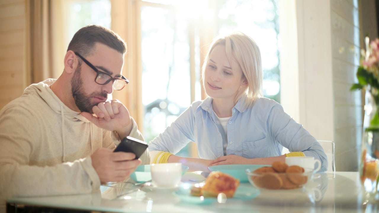 A man with glasses looks at a phone while a woman watches him at a breakfast table.