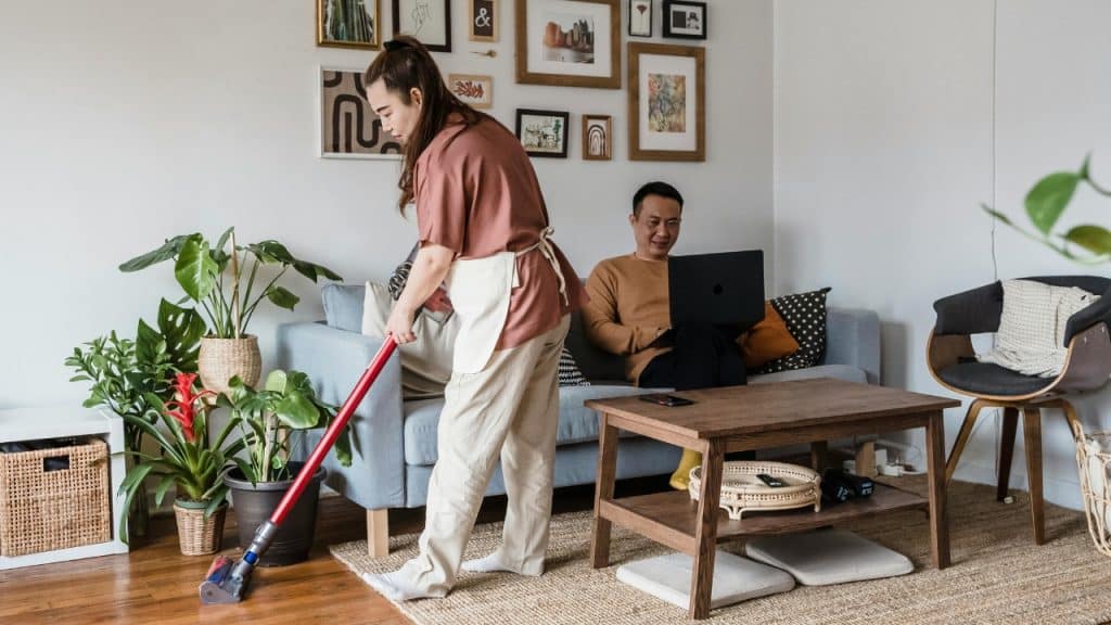 A woman vacuums a living room while a man sits on a sofa using a laptop.