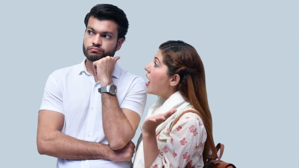 A woman gestures and speaks toward a man who is looking away with a hand on his chin.