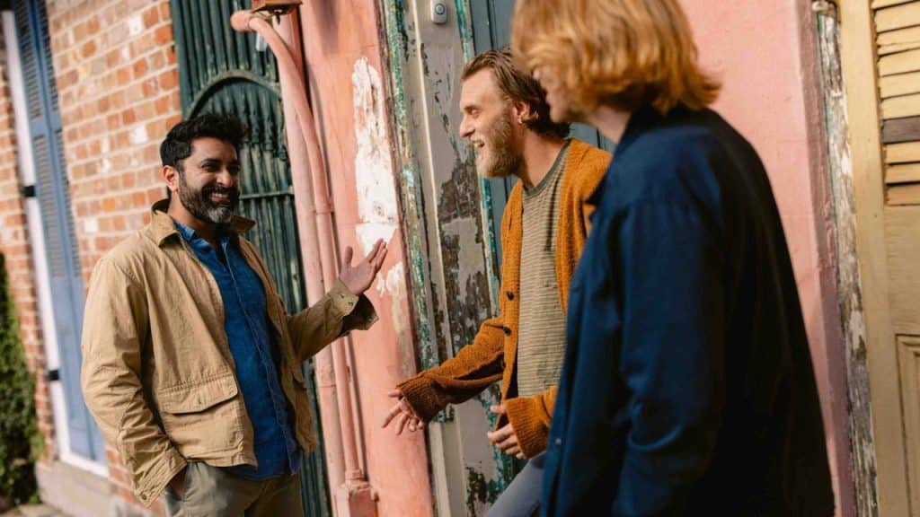 Three men stand on a sidewalk outside a brick building, talking and laughing together.