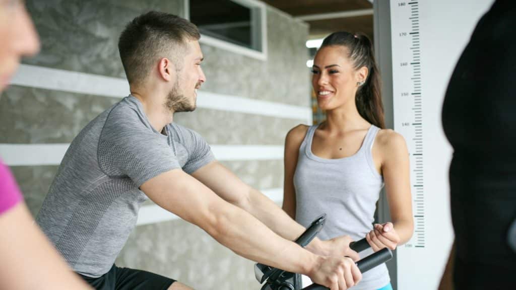 A man on an exercise bike looks up and smiles at a woman standing in a gym.