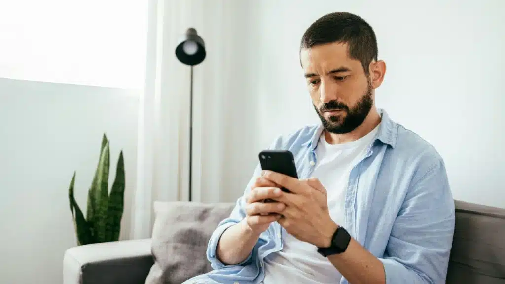 A bearded man in a light blue shirt sits on a sofa using a smartphone.
