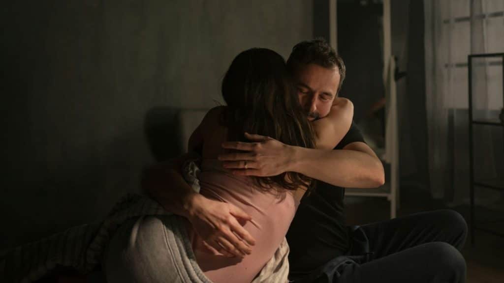 A man and woman embrace closely while sitting on a bed in a dimly lit room.