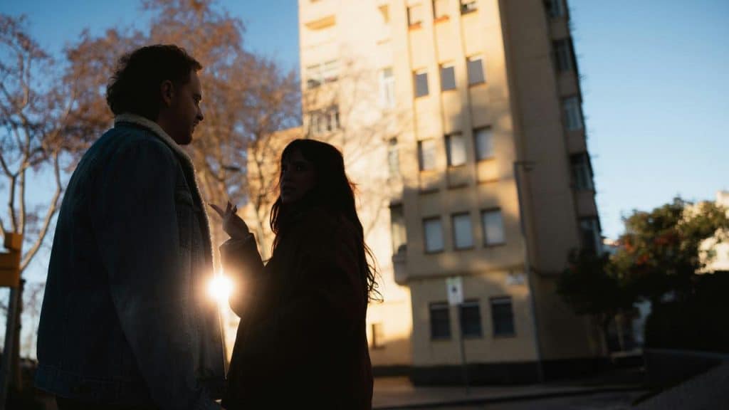 A man and woman standing outside, talking in the sunlight.