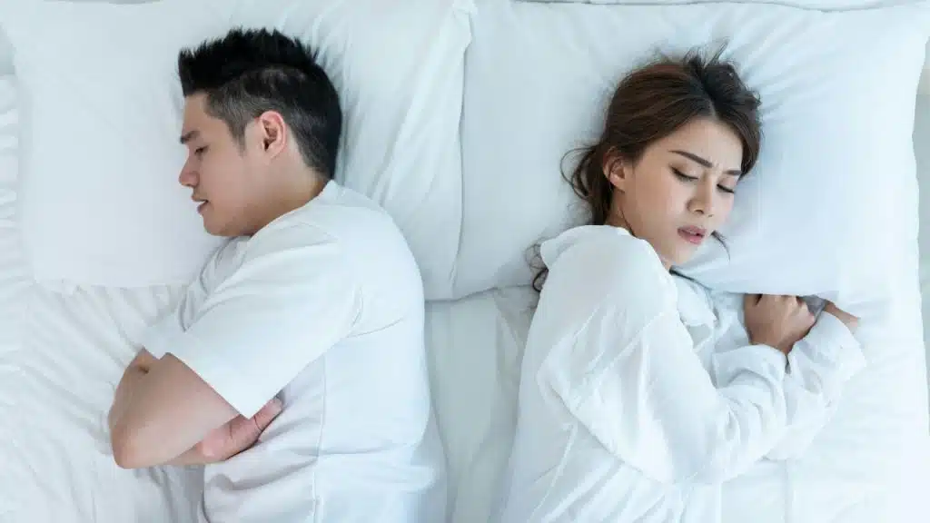 A man and woman lie back-to-back in a bed with white sheets, appearing distressed or upset.