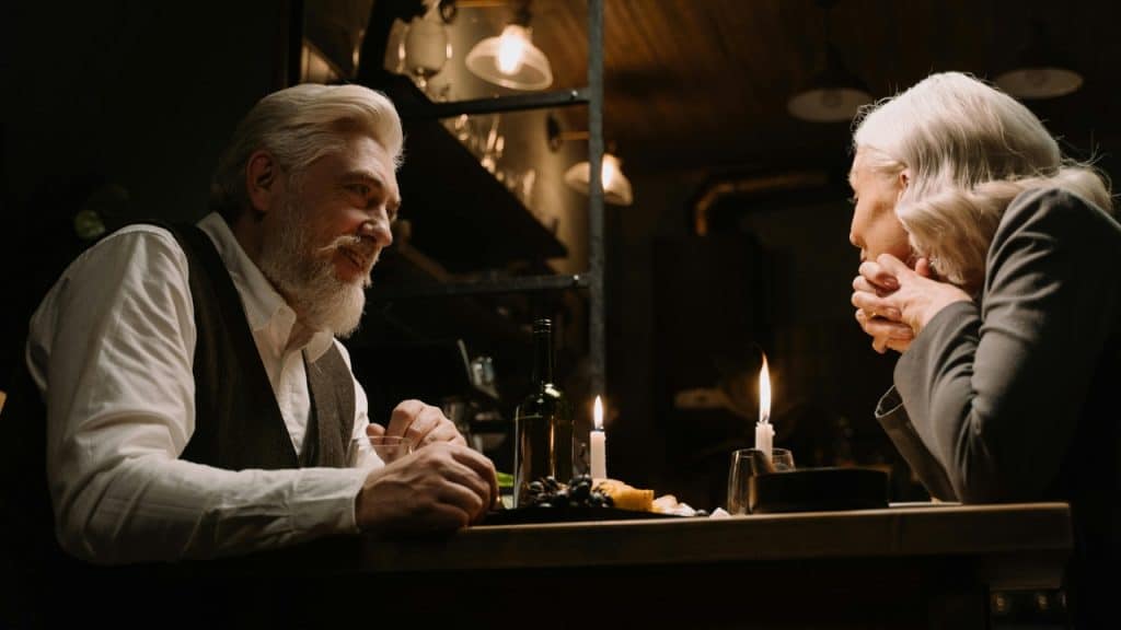 An older man and woman sit at a candlelit table with wine and fruit snacks.