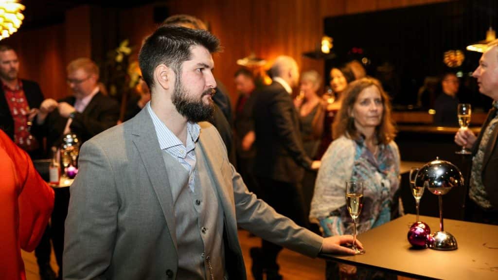 A bearded man in a grey suit stands at a crowded event near a table.