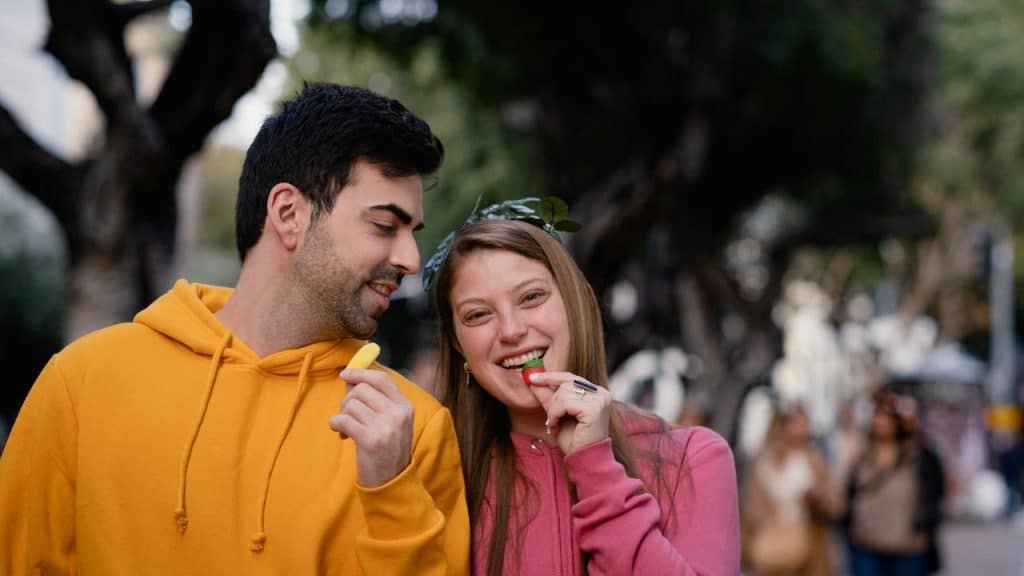 A man and woman smiling while eating candy outdoors.