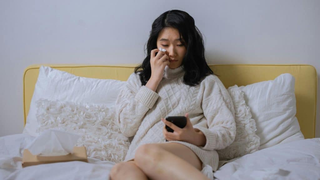A woman in a white sweater sits on a bed holding a phone and a tissue.