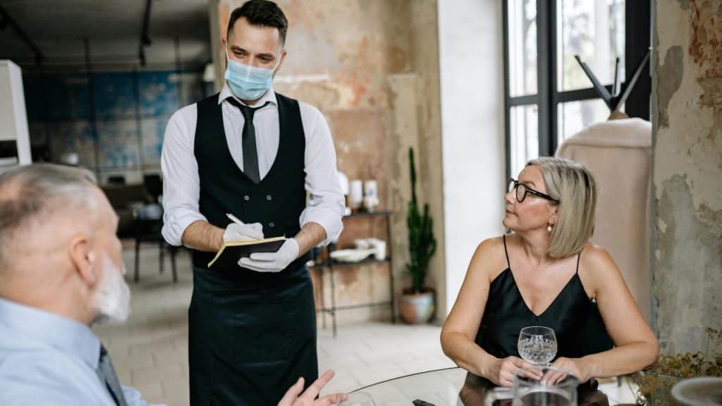 A server in a mask and vest takes an order from a couple at a table.