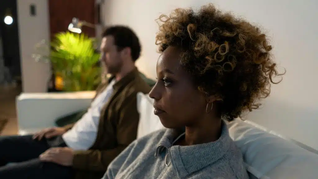 A woman with curly hair looks ahead while a man sits blurred in the background.