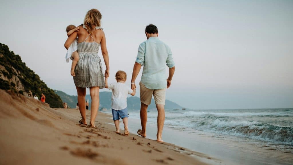 A family of four walks away from the camera on a sandy beach near waves.
