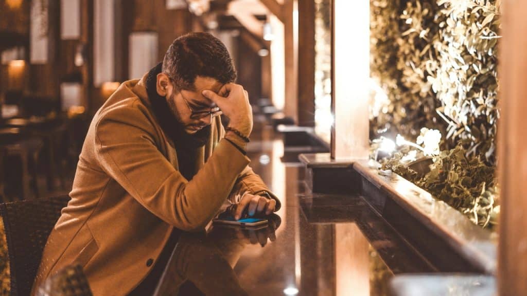 A man in a tan coat sitting at a bar looking down at a smartphone.