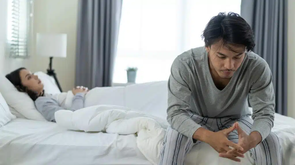 A distressed man sits on the edge of a bed in a bright bedroom, while a woman lies asleep or unresponsive behind him.