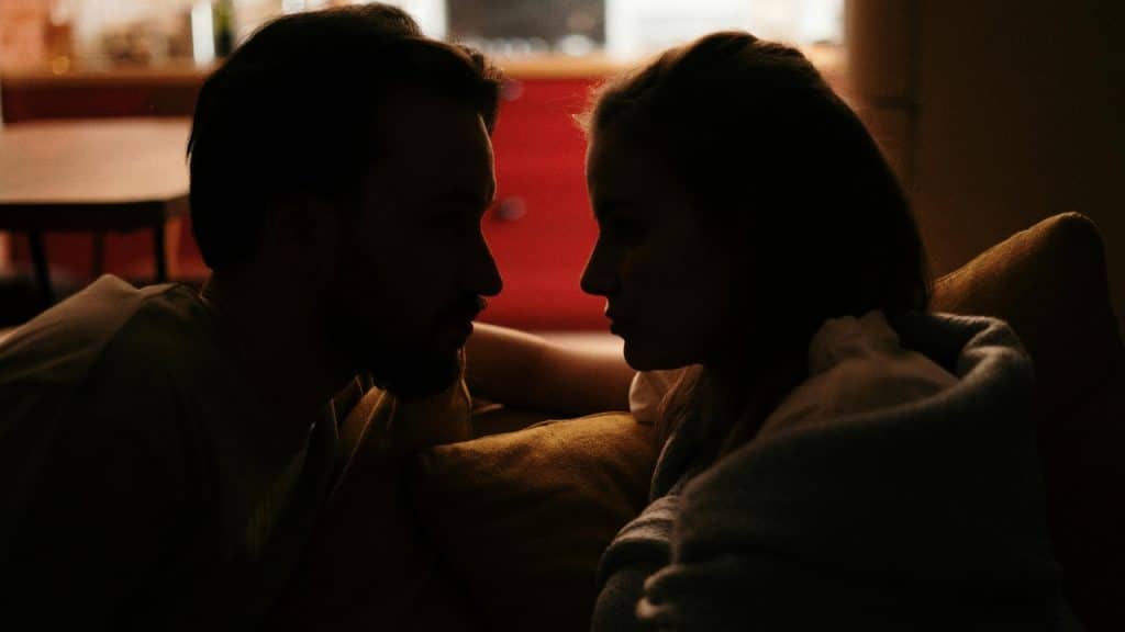 A man and woman look at each other in a dimly lit room with shadows.