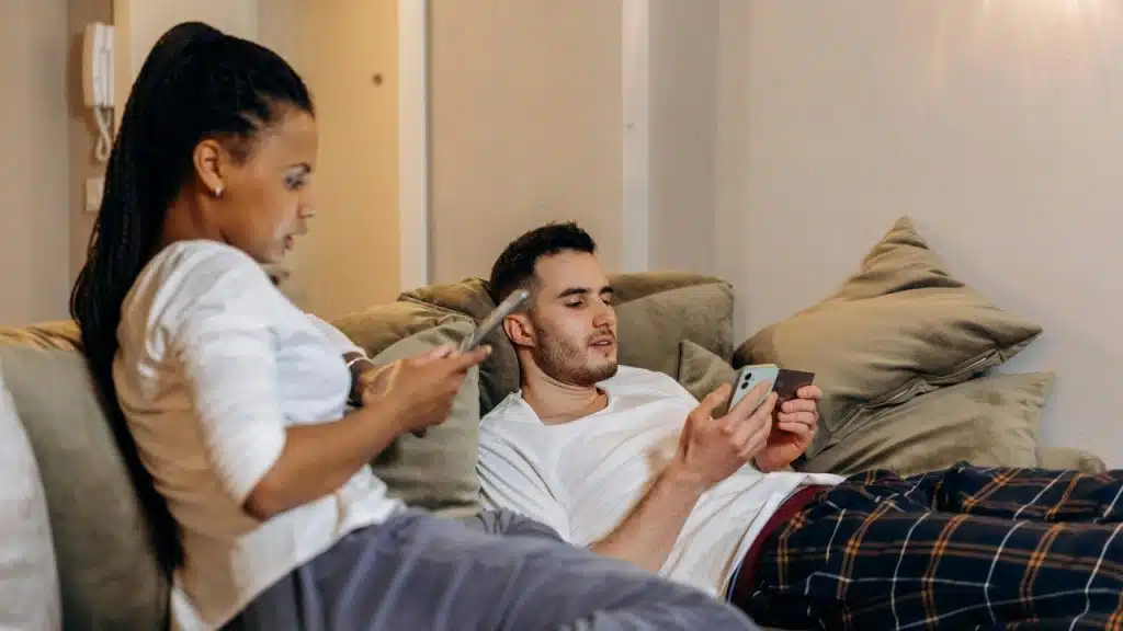 Two people sit on a couch together while looking at their own separate mobile phone screens.