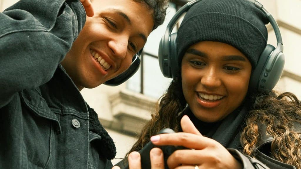 A pair of friends wearing headphones and smiling at a phone.