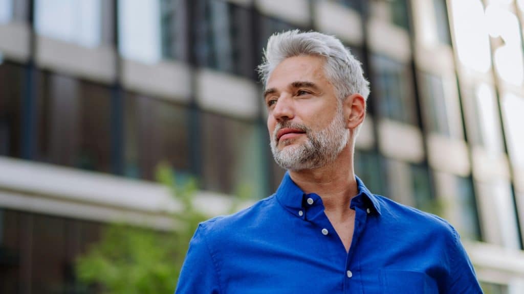 A man with gray hair and a blue button-down shirt looks toward the side near city buildings.