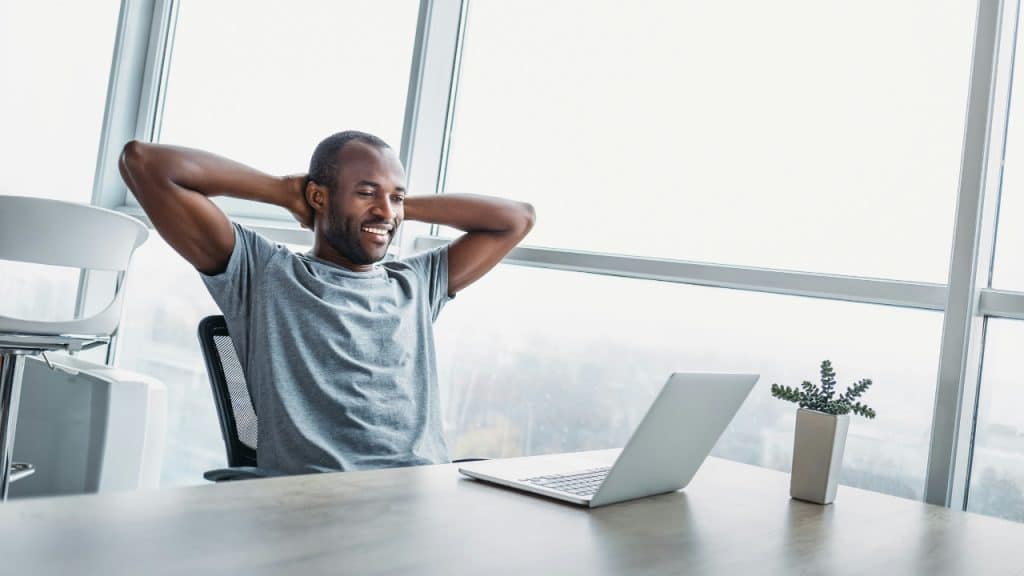A man sits at a wooden desk with a laptop, leaning back with hands behind head.