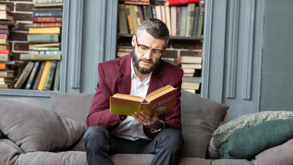 A bearded man in a maroon blazer sits on a couch reading a book.