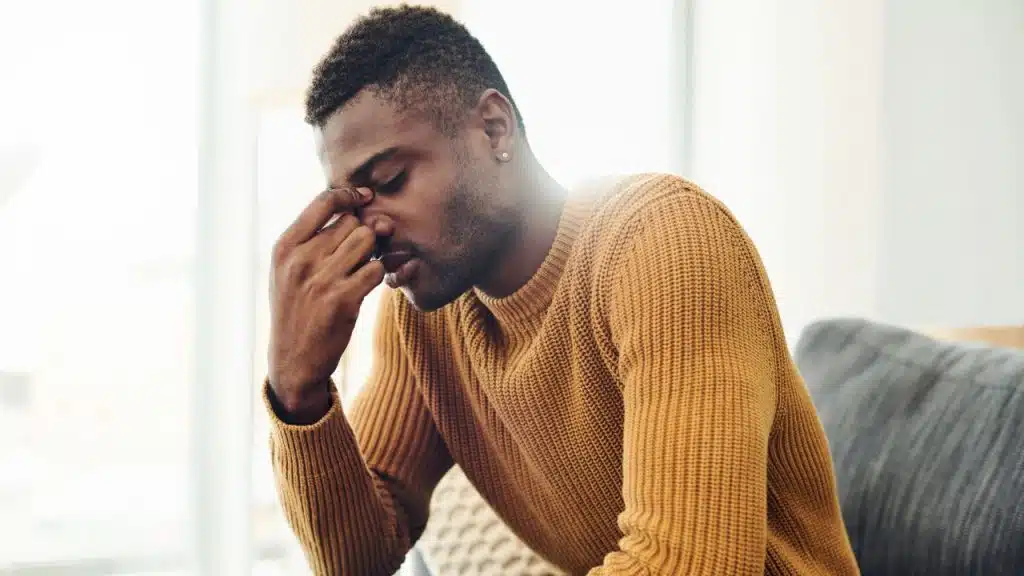A man in a mustard yellow sweater sits down while pinching the bridge of his nose.