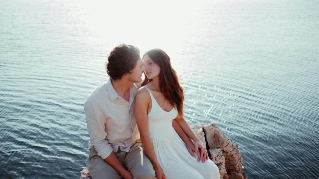 A couple sitting close together by the water at sunset.