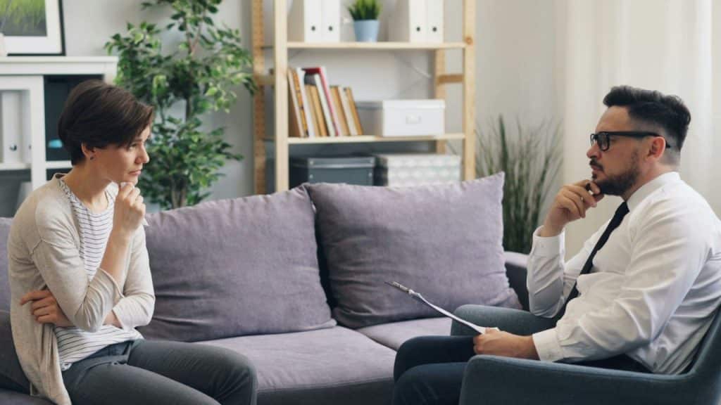 A woman talks to a man holding a clipboard while sitting on a couch.