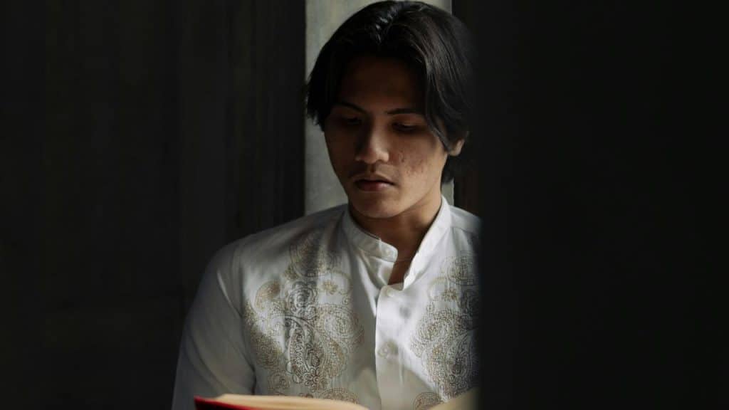 A man in a white embroidered shirt reading a book in dim light.