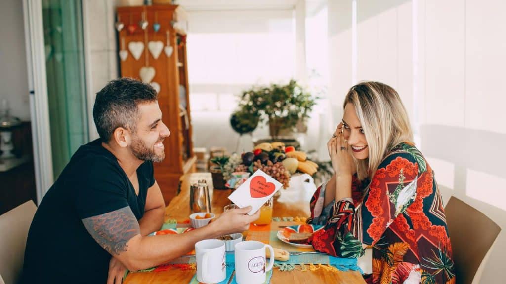 A man giving a card to a smiling woman while sitting together at a breakfast table.