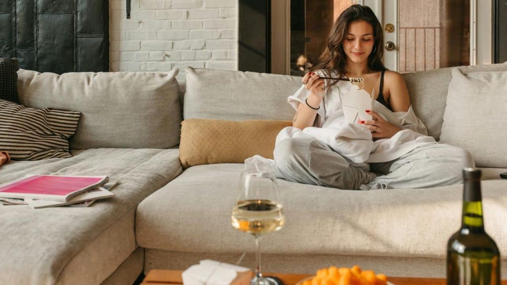 A woman relaxing on her couch eating takeout food with wine and snacks on the table.