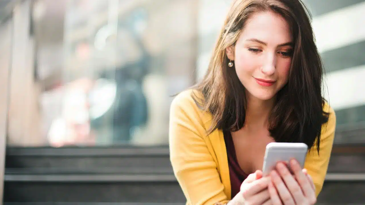 A woman in a yellow cardigan looking at her smartphone indoors.