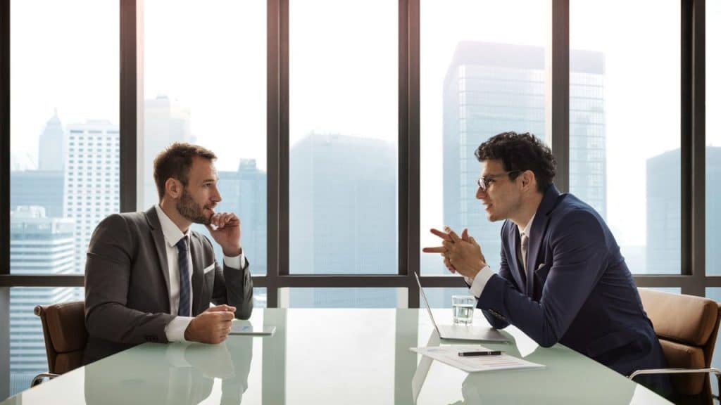 Two men in suits sitting at a table across from each other in a city office.
