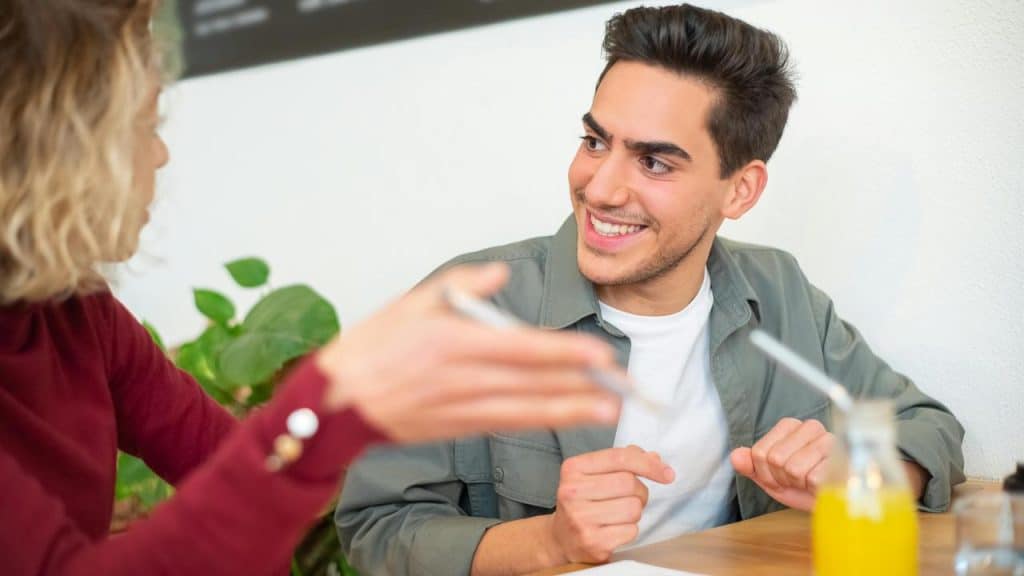 A man smiling while talking with a woman at a table with a drink nearby.