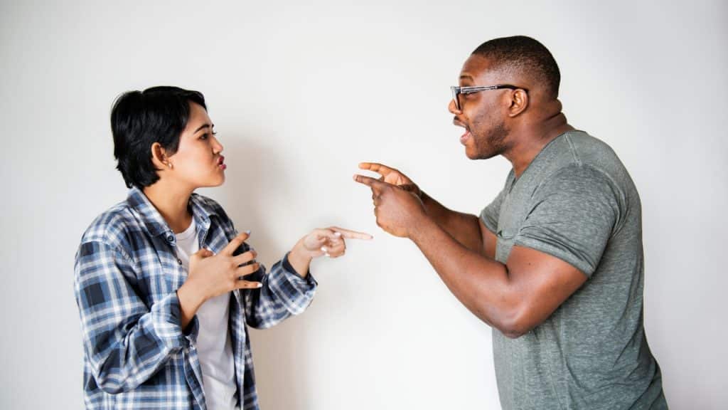 A woman in a flannel shirt and a man in glasses point toward each other.